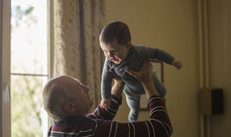 man wearing maroon, white, and blue stripe long-sleeved shirt lifting up baby wearing gray onesie