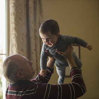 man wearing maroon, white, and blue stripe long-sleeved shirt lifting up baby wearing gray onesie