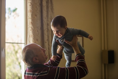 man wearing maroon, white, and blue stripe long-sleeved shirt lifting up baby wearing gray onesie grandparents teams background