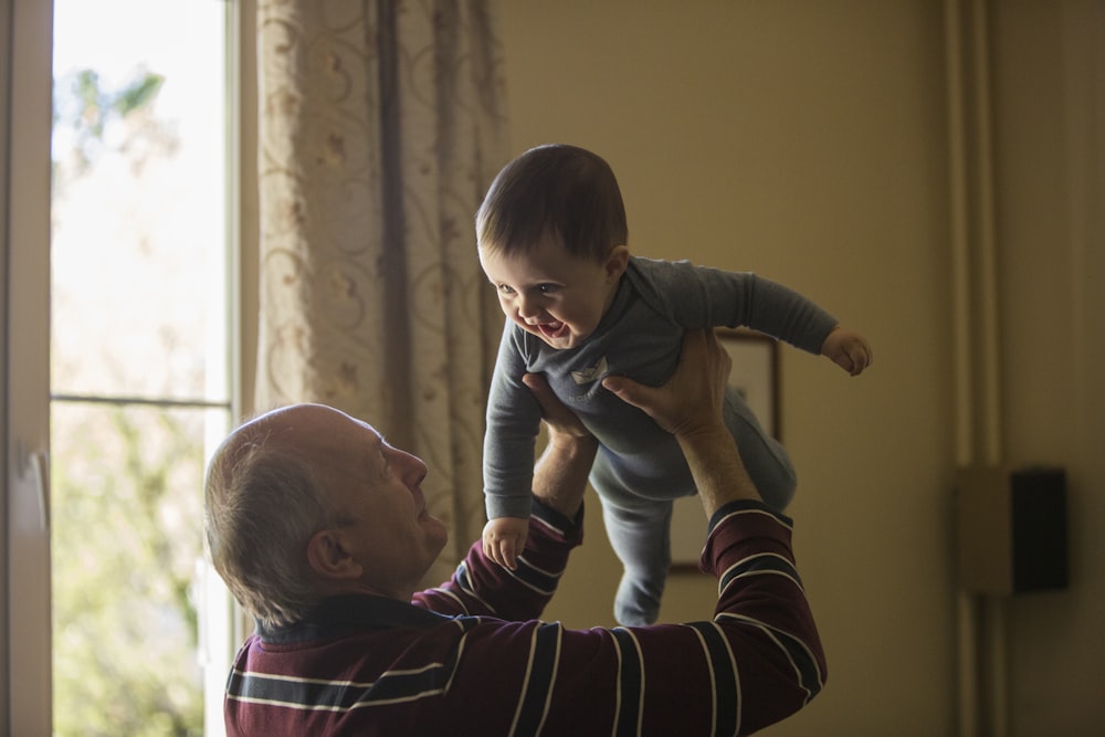 man wearing maroon, white, and blue stripe long-sleeved shirt lifting up baby wearing gray onesie