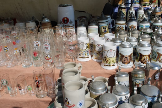 A collection of various glass and ceramic beer steins and mugs, some with ornate designs or logos, are displayed on a table. The collection includes both tall glass beer mugs and traditional German-style steins with metal lids.