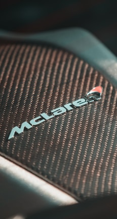 Close-up of a section of a vehicle featuring a textured, carbon-fiber-like surface with the McLaren logo prominently displayed, suggesting performance and luxury.