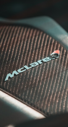 Close-up of a section of a vehicle featuring a textured, carbon-fiber-like surface with the McLaren logo prominently displayed, suggesting performance and luxury.