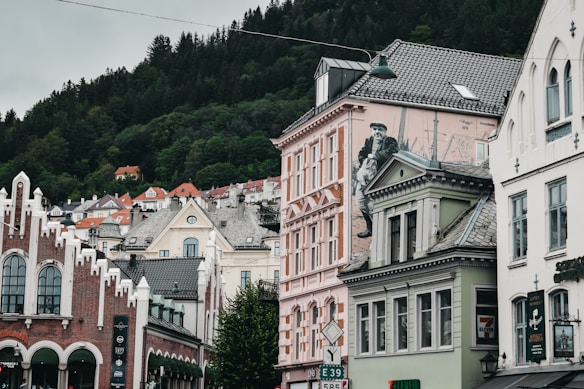 A street view in a European town with colorful buildings featuring distinct architectural styles. A prominent mural of a person is displayed on one of the buildings. In the background, lush, forested hills create a natural contrast to the urban setting. The buildings have a variety of pastel hues, ornate facades, and large windows.