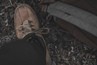 A worn brown hiking boot is laced up and resting on a grassy ground with patches of dry leaves. Next to the boot, a gray strap from a backpack is visible.