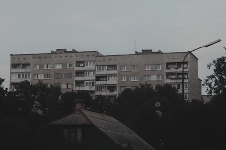 A large, rectangular, concrete apartment building with multiple floors is seen surrounded by foliage. The building's facade is plain and utilitarian, featuring rows of windows and some balconies with clothes hanging. The foreground includes the roof of a smaller structure, and the sky is overcast.