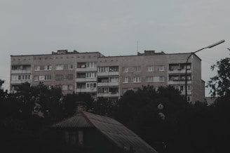 A large, rectangular, concrete apartment building with multiple floors is seen surrounded by foliage. The building's facade is plain and utilitarian, featuring rows of windows and some balconies with clothes hanging. The foreground includes the roof of a smaller structure, and the sky is overcast.