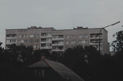 A large, rectangular, concrete apartment building with multiple floors is seen surrounded by foliage. The building's facade is plain and utilitarian, featuring rows of windows and some balconies with clothes hanging. The foreground includes the roof of a smaller structure, and the sky is overcast.