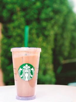 A clear plastic cup filled with a light brown iced beverage featuring the Starbucks logo sits on a white surface. In the background, there is a lush, green, out-of-focus hedge.