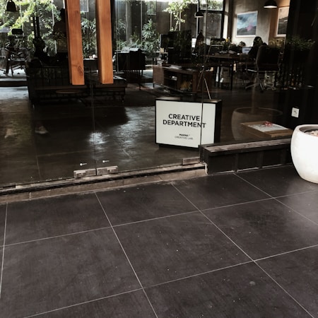 A modern office setting with glass doors leading to an interior workspace. Inside, people are working at desks with computers, surrounded by indoor plants and framed artwork on the walls. A sign reading 'Creative Department' is prominently displayed.