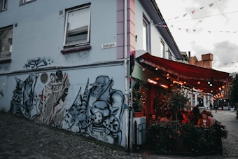 A street corner featuring a detailed wall mural with fantasy-themed characters, including knights and mythical creatures, painted in grayscale. Adjacent to the mural is a bustling outdoor caf&eacute; with people seated under a warm, red canopy. The cobblestone street adds a quaint charm, and festive pennants hang overhead.