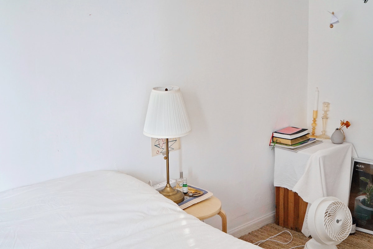 clean minimalist small apartment bedroom with no headboard, white walls, lamp, and books on side table