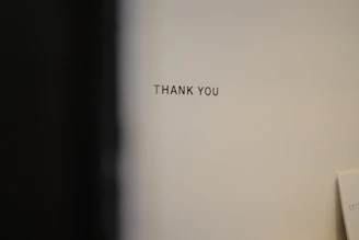 A plain wall with the words 'THANK YOU' printed in black uppercase letters. The background is a neutral color, and there is a slight shadow or dark area on the left side.