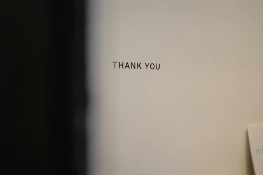 A plain wall with the words 'THANK YOU' printed in black uppercase letters. The background is a neutral color, and there is a slight shadow or dark area on the left side.