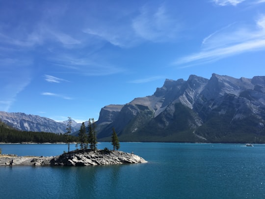 body of water near black mountains under blue sky at daytime in Lake Minnewanka Trail Canada