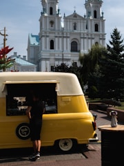 A person stands in front of a bright yellow and white vintage van repurposed as a food truck. The vehicle is parked on a cobbled street with a large, ornate church featuring tall towers with clocks in the background. A small table with a white vase containing colorful flowers is to the side. The scene is set in a sunny outdoor area with trees and decorative elements.