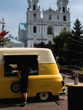 A person stands in front of a bright yellow and white vintage van repurposed as a food truck. The vehicle is parked on a cobbled street with a large, ornate church featuring tall towers with clocks in the background. A small table with a white vase containing colorful flowers is to the side. The scene is set in a sunny outdoor area with trees and decorative elements.