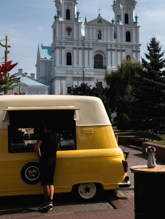 A person stands in front of a bright yellow and white vintage van repurposed as a food truck. The vehicle is parked on a cobbled street with a large, ornate church featuring tall towers with clocks in the background. A small table with a white vase containing colorful flowers is to the side. The scene is set in a sunny outdoor area with trees and decorative elements.