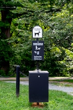 A pet waste station is situated in a park-like environment. The station includes a waste bin with a sign above it requesting visitors to clean up after their pets. The area is surrounded by lush green grass and trees, creating a natural setting.