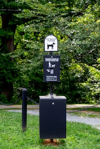 A pet waste station is situated in a park-like environment. The station includes a waste bin with a sign above it requesting visitors to clean up after their pets. The area is surrounded by lush green grass and trees, creating a natural setting.