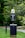A pet waste station is situated in a park-like environment. The station includes a waste bin with a sign above it requesting visitors to clean up after their pets. The area is surrounded by lush green grass and trees, creating a natural setting.