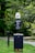 A pet waste station is situated in a park-like environment. The station includes a waste bin with a sign above it requesting visitors to clean up after their pets. The area is surrounded by lush green grass and trees, creating a natural setting.