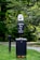 A pet waste station is situated in a park-like environment. The station includes a waste bin with a sign above it requesting visitors to clean up after their pets. The area is surrounded by lush green grass and trees, creating a natural setting.