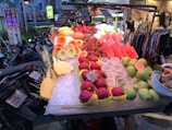 An outdoor market stall displays a variety of fresh, colorful fruits including cut pineapples, watermelons, dragon fruits, and guavas. The fruits are neatly arranged on ice, with some already prepared and others whole. Bicycles and motorbikes are parked along the side, and a boutique with clothing can be seen in the background.