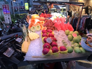 An outdoor market stall displays a variety of fresh, colorful fruits including cut pineapples, watermelons, dragon fruits, and guavas. The fruits are neatly arranged on ice, with some already prepared and others whole. Bicycles and motorbikes are parked along the side, and a boutique with clothing can be seen in the background.