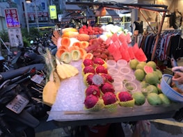 An outdoor market stall displays a variety of fresh, colorful fruits including cut pineapples, watermelons, dragon fruits, and guavas. The fruits are neatly arranged on ice, with some already prepared and others whole. Bicycles and motorbikes are parked along the side, and a boutique with clothing can be seen in the background.