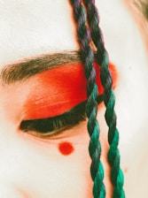A close-up of an eye adorned with bright red eyeshadow and a red dot below it. The eyebrows are dark and defined. A colorful braid, mixing blue and purple hues, is draped across the face.