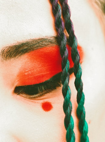 A close-up of an eye adorned with bright red eyeshadow and a red dot below it. The eyebrows are dark and defined. A colorful braid, mixing blue and purple hues, is draped across the face.