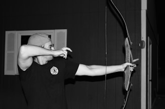 A person is practicing archery, wearing a cap and glasses, aiming an arrow with a traditional bow. The background features a building with windows.