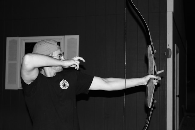 A person is practicing archery, wearing a cap and glasses, aiming an arrow with a traditional bow. The background features a building with windows.
