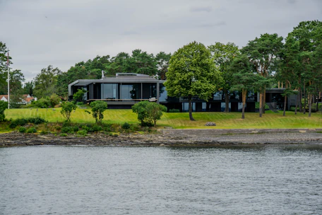 A stunning modern luxury home with large glass windows overlooking a serene waterfront at sunset.