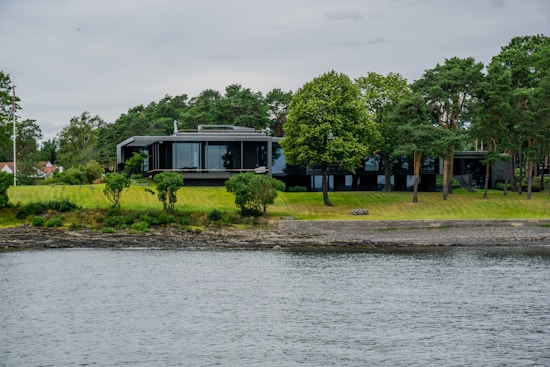 A modern house with large glass windows is surrounded by lush green trees and a well-maintained lawn. The house is located by the water, creating a serene and tranquil atmosphere.