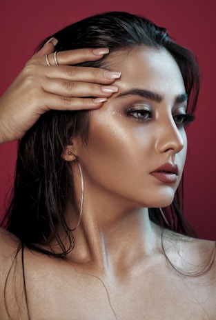 A woman with long dark hair styled back, displaying a side profile. Her skin appears smooth with a glowing, dewy look, and she wears makeup with bold eyebrows, smoky eyeshadow, and dark red lipstick. She is holding her left hand over her forehead with manicured nails and wearing a pair of hoop earrings. The background is a solid deep red color.