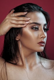 A woman with long dark hair styled back, displaying a side profile. Her skin appears smooth with a glowing, dewy look, and she wears makeup with bold eyebrows, smoky eyeshadow, and dark red lipstick. She is holding her left hand over her forehead with manicured nails and wearing a pair of hoop earrings. The background is a solid deep red color.