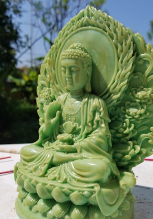 A jade-colored Buddha statue intricately carved with detailed patterns is seated in a meditative pose on a lotus base. Surrounding the statue is a lush garden with a clear blue sky providing a serene backdrop.