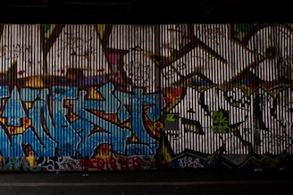 A corrugated metal wall is covered in vibrant and intricate graffiti art. The artwork includes several layers of bright colors, such as blue, red, and green, with bold lettering and abstract shapes interspersed with various tags.