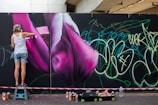 A person is spray painting a large mural on a wall featuring vibrant pink and purple flower-like shapes. The person is wearing casual clothing and standing on a small step stool. A variety of spray paint cans are scattered on the ground nearby. The wall is also covered with green and yellow graffiti-style writing.