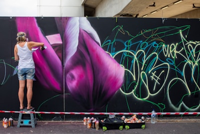 A person is spray painting a large mural on a wall featuring vibrant pink and purple flower-like shapes. The person is wearing casual clothing and standing on a small step stool. A variety of spray paint cans are scattered on the ground nearby. The wall is also covered with green and yellow graffiti-style writing.
