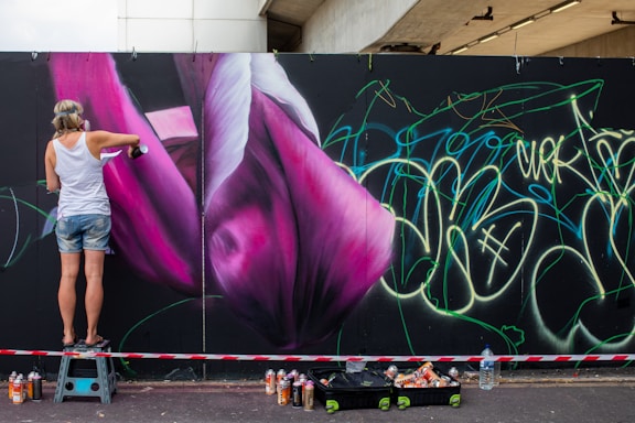 A person is spray painting a large mural on a wall featuring vibrant pink and purple flower-like shapes. The person is wearing casual clothing and standing on a small step stool. A variety of spray paint cans are scattered on the ground nearby. The wall is also covered with green and yellow graffiti-style writing.