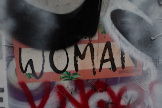 A piece of street art with the word 'WOMAN' prominently displayed in bold, black letters against a multicolored background. The letters have a hand-painted style with a few green leaf motifs around them. The background shows layers of graffiti, including shades of gray and red, and appears textured. The image includes the handle '@IVYROSELONDON' in smaller text.