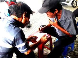 Two men are sitting on small stools on a paved area, intently focusing on a piece of paper. One man is writing while the other appears to be explaining or contemplating, with a hand on his chin. A motorcycle is parked nearby in the background.