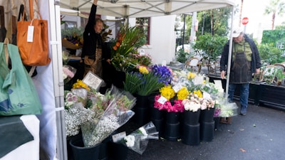 A vibrant outdoor flower market stall displayed under a white canopy features a variety of colorful flowers, organized in black buckets. Two individuals, likely vendors, are present; one is adjusting the flowers while the other seems to be taking payment. Signs indicating flower prices are visible. Bags hang on the side of the stall and some greenery is visible in the background.