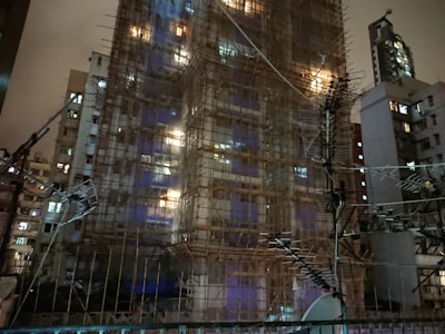 A building under construction is covered with scaffolding and surrounded by various antennas. The structure is lit from within, casting a warm glow against the dark, cloudy sky. Nearby buildings are visible in the background, contributing to an urban nighttime setting.