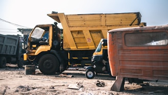 A large yellow dump truck is parked in an outdoor setting surrounded by dirt and debris. A small rickshaw is visible in front of the truck, alongside an old, rusted vehicle. The scene appears to be in a scrapyard or industrial area with other vehicles in the background.