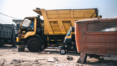 A large yellow dump truck is parked in an outdoor setting surrounded by dirt and debris. A small rickshaw is visible in front of the truck, alongside an old, rusted vehicle. The scene appears to be in a scrapyard or industrial area with other vehicles in the background.
