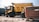 A large yellow dump truck is parked in an outdoor setting surrounded by dirt and debris. A small rickshaw is visible in front of the truck, alongside an old, rusted vehicle. The scene appears to be in a scrapyard or industrial area with other vehicles in the background.